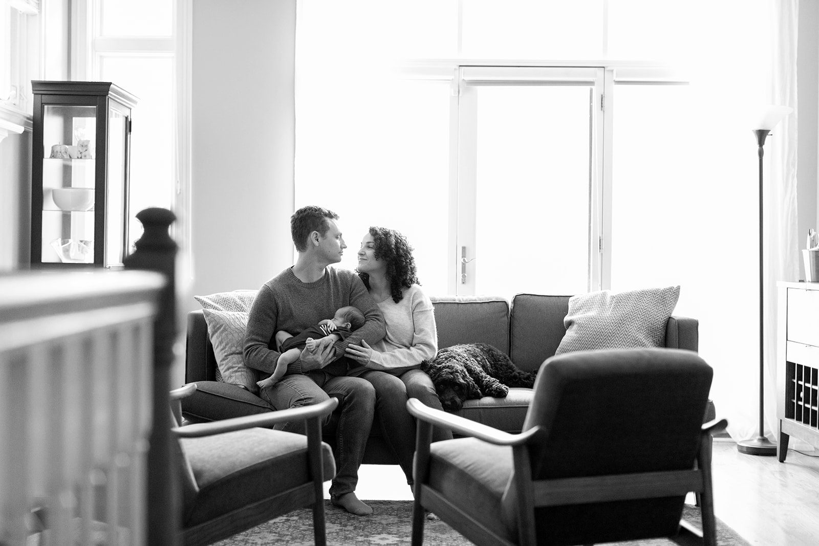man and woman smile at each other while sitting on couch holding newborn baby