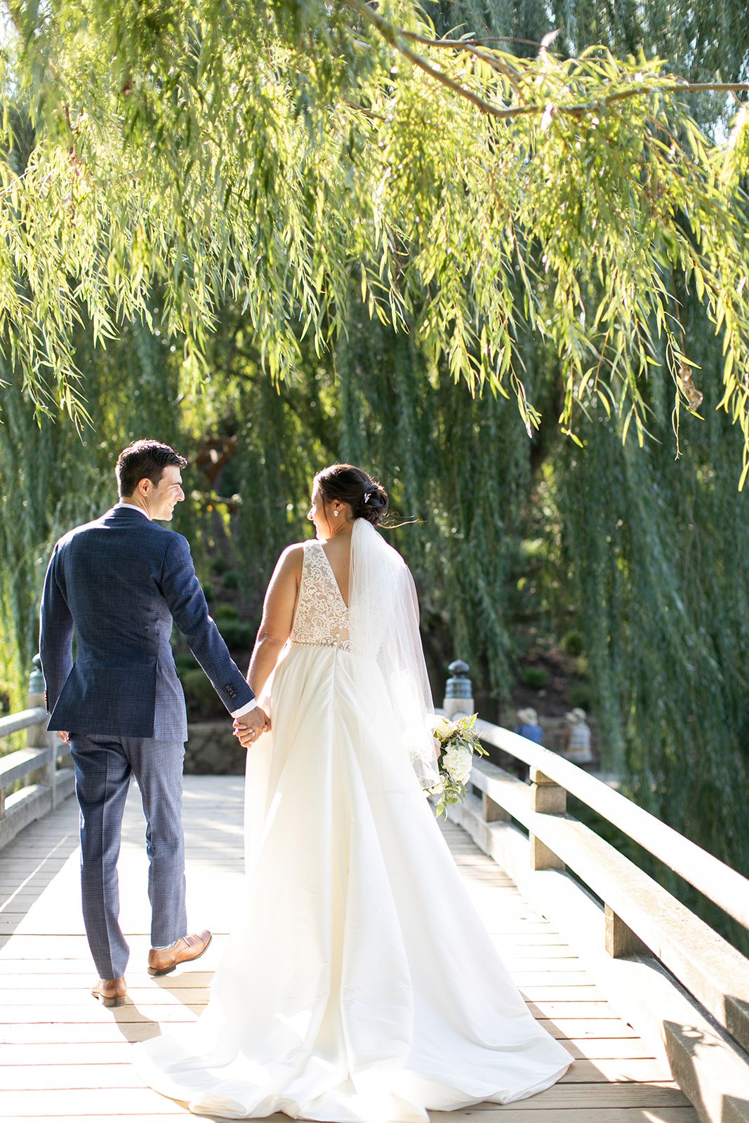 Bride and groom cross bridge surrounded by willow trees at Chicago Botanic Garden